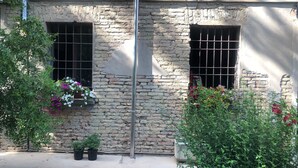 A rustic building with two barred windows and a metal pole in the center. There are potted plants and flowers around the building. - Family room Fico's colors (San Lorenzo)