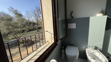 Ein Badezimmer mit Toilette, Waschbecken und Spiegel. Blick auf Bäume und ein Balkon.