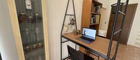 A room with a wooden desk, a laptop, a black chair, a bookshelf, and a glass cabinet with wine bottles.