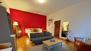 Ein modernes Wohnzimmer mit einer roten Akzentwand, einem grauen Sofa, einem Holztisch und Blick in das angrenzende Schlafzimmer.