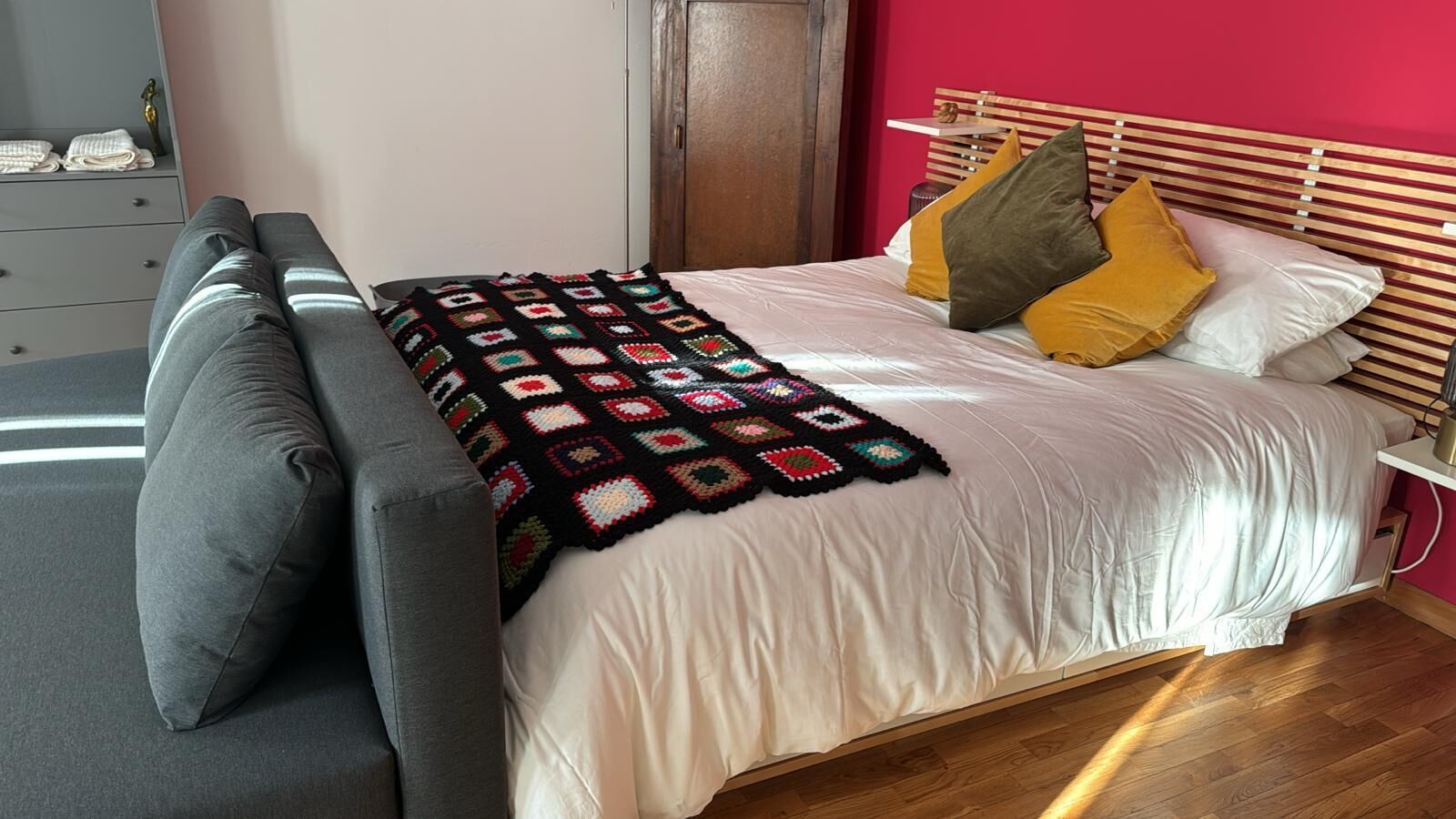A neatly made bed with a colorful blanket, a grey sofa, and a wooden headboard.