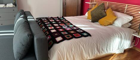 A neatly made bed with a colorful blanket, a grey sofa, and a wooden headboard.