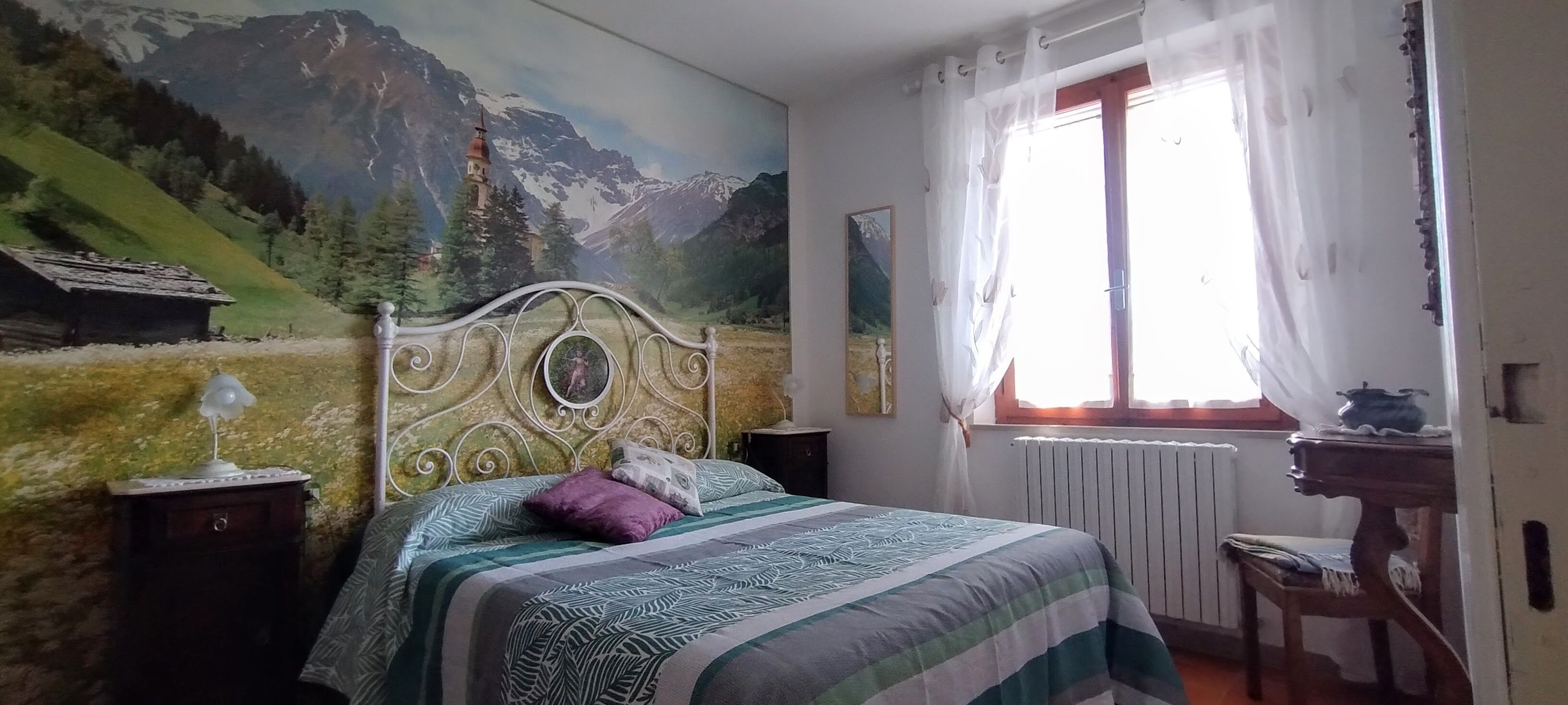 Un dormitorio con un mural de paisaje montañoso, una cama con ropa de cama estampada, una mesita de noche con una lámpara y una ventana con cortinas.