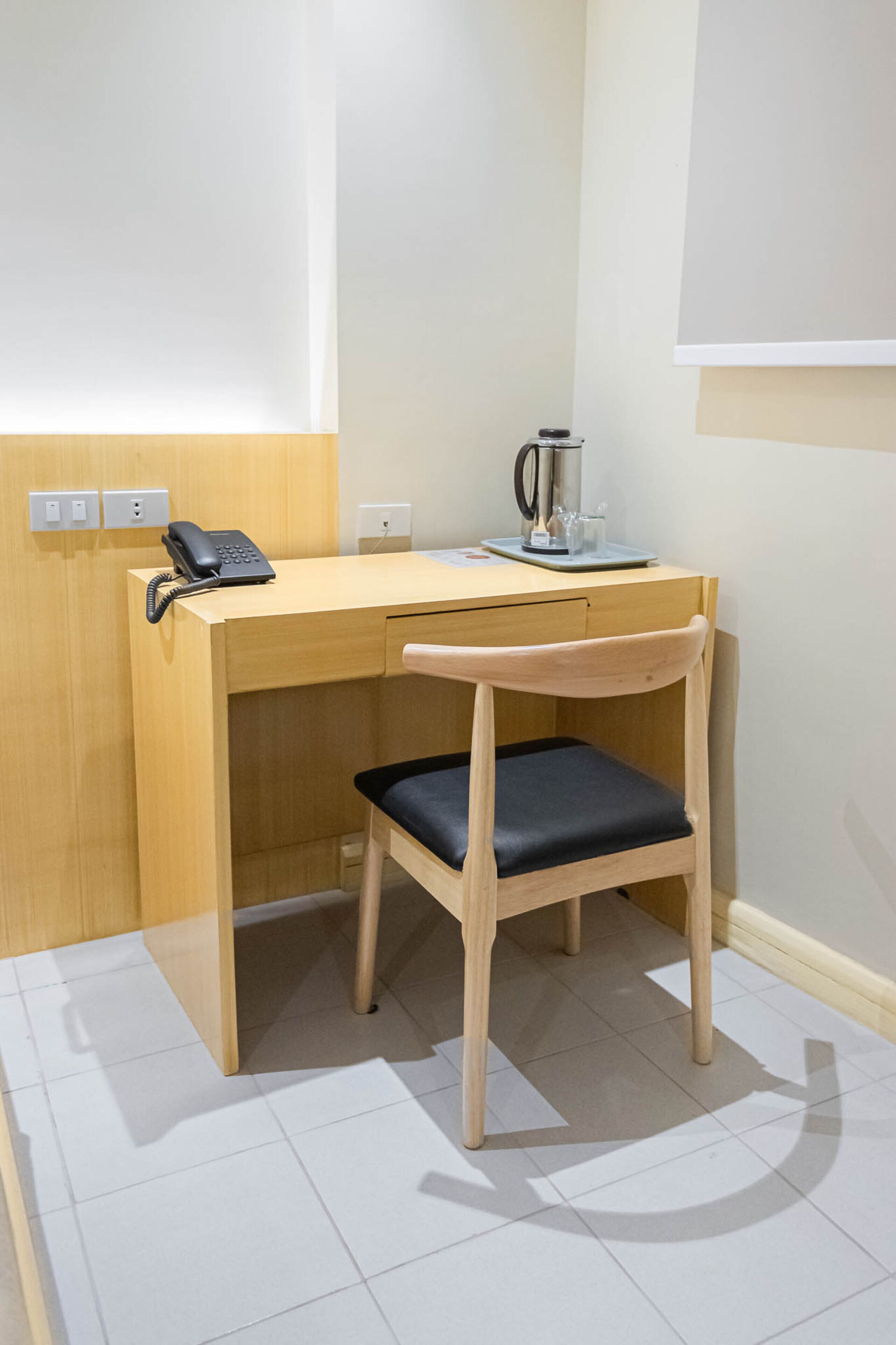 A wooden desk with a telephone, a coffee machine, and a chair with a black cushion.