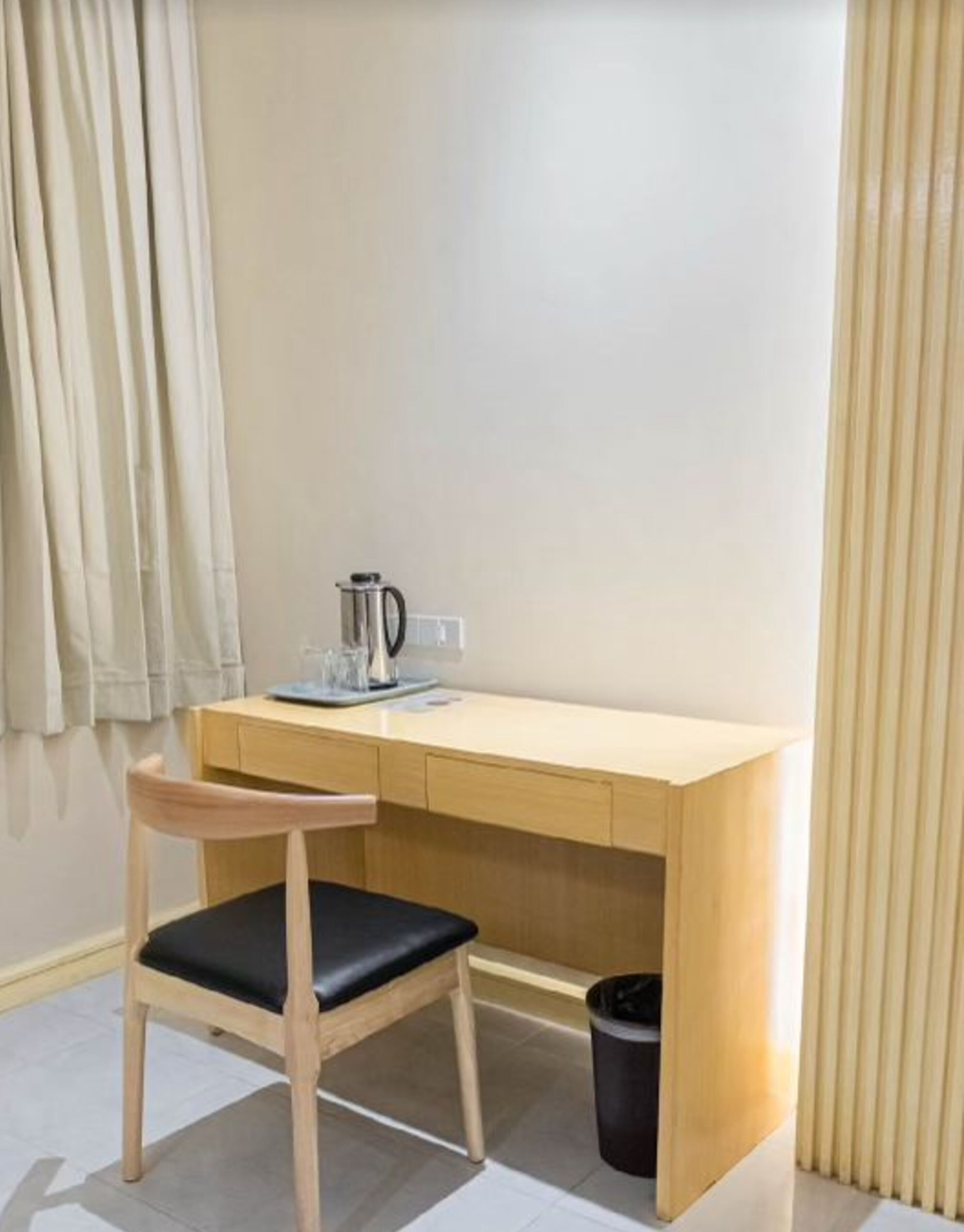 Una habitación pequeña y sencilla con un escritorio de madera, una silla, una cafetera y un cubo de basura.