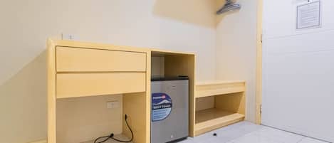 A compact room with a wooden desk, a small refrigerator, and a light shelf.