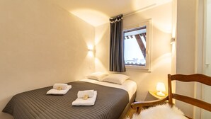Une chambre d’hôtel avec un lit, des oreillers, une table de chevet avec une lampe, une chaise avec un coussin moelleux et une fenêtre avec des rideaux.