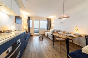 Moderni keittiö, jossa on sisäänrakennettu uuni, ruokailutila puupöydällä ja -tuoleilla, sohva ja suuri ikkuna, jossa on verhot.