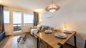 Un coin repas avec une table en bois dressée pour deux personnes, un luminaire suspendu et une vue sur le balcon par la fenêtre.