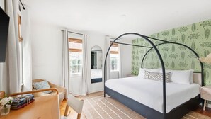 A bedroom with a four-poster bed, a wooden desk with books, a wicker chair, and a view of a bathroom through an open door. - The OakTree | DT Charleston Gem - Near King Street (Charleston)