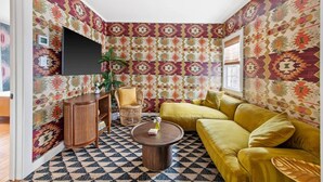 A corner living room with patterned wallpaper, a yellow sofa, a round coffee table, and a TV mounted on the wall. - The OakTree | Bohemian Charm in DT Charleston! (Charleston)