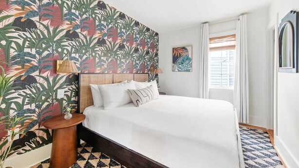 A bedroom with a bed, a nightstand, a lamp, a window with blinds, and a tropical wallpaper. - The OakTree | Bohemian Charm in DT Charleston! (Charleston)