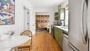 A narrow kitchen with green cabinets, a white refrigerator, and a dining area with a white table and wicker chairs. - The OakTree | Bohemian Charm in DT Charleston! (Charleston)
