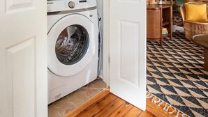 A laundry room with stacked washing machines, a patterned rug, and a wicker chair. - The OakTree | Bohemian Charm in DT Charleston! (Charleston)
