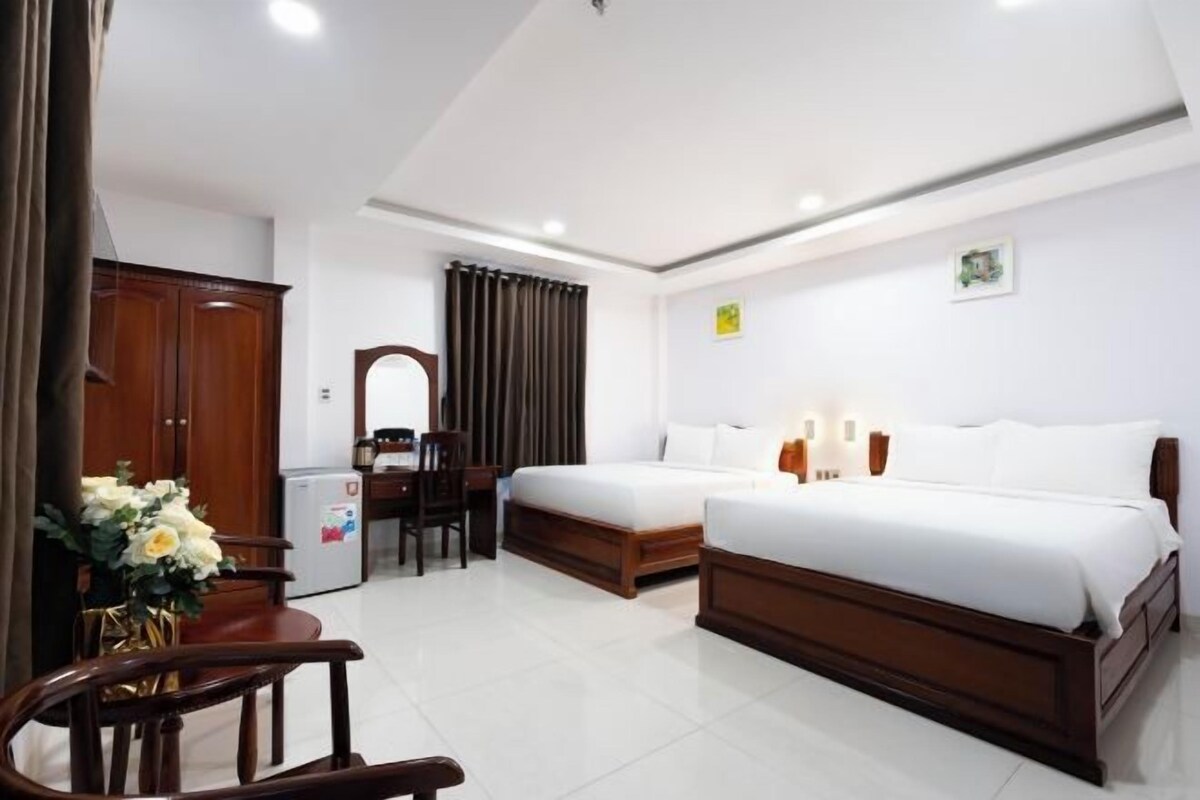 A hotel room with two beds, a desk, a chair, a refrigerator, and a small table with a vase of flowers.