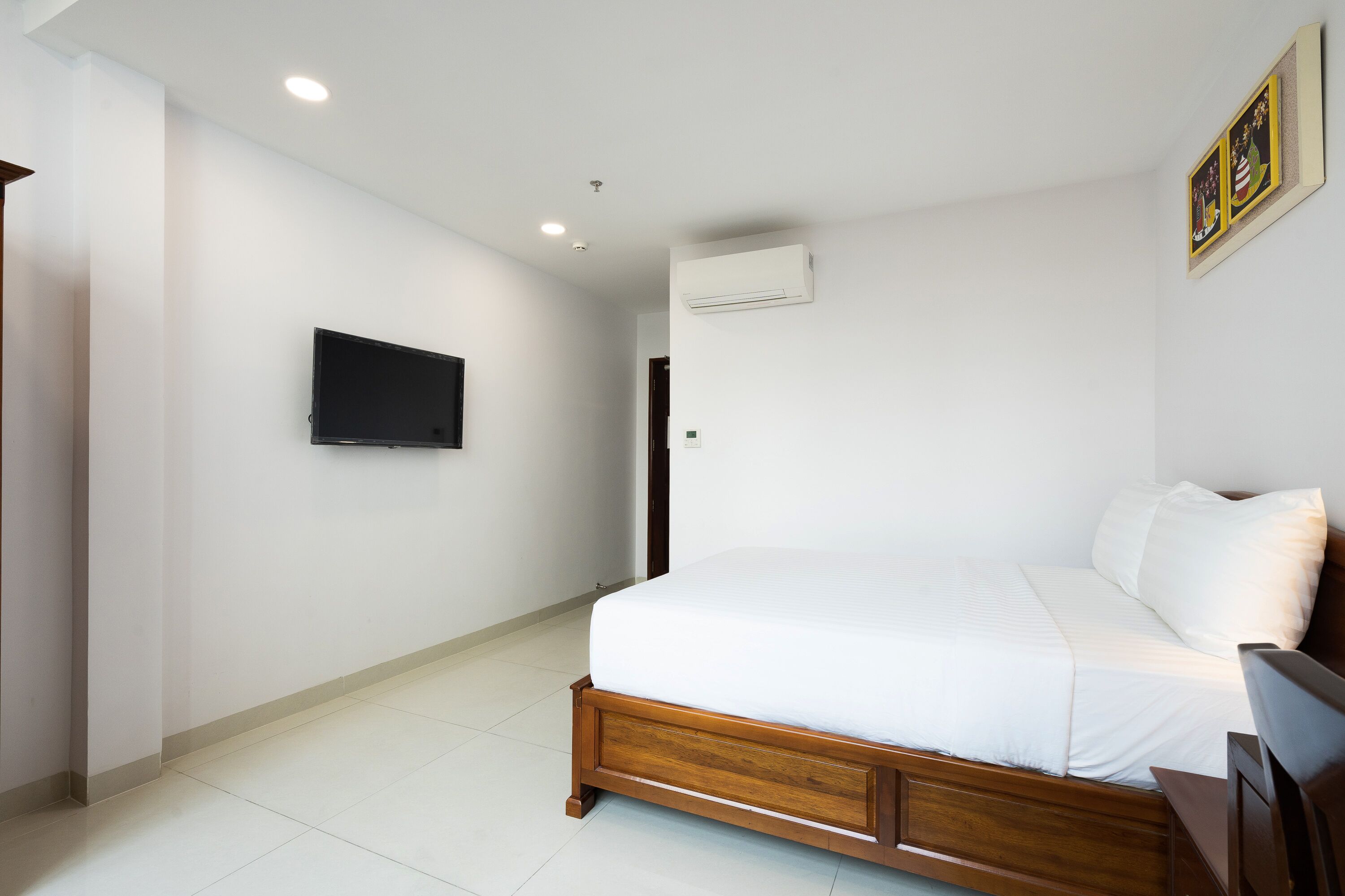 A bedroom with a wooden bed, white bedding, a mounted TV, and an air conditioning unit on the wall.