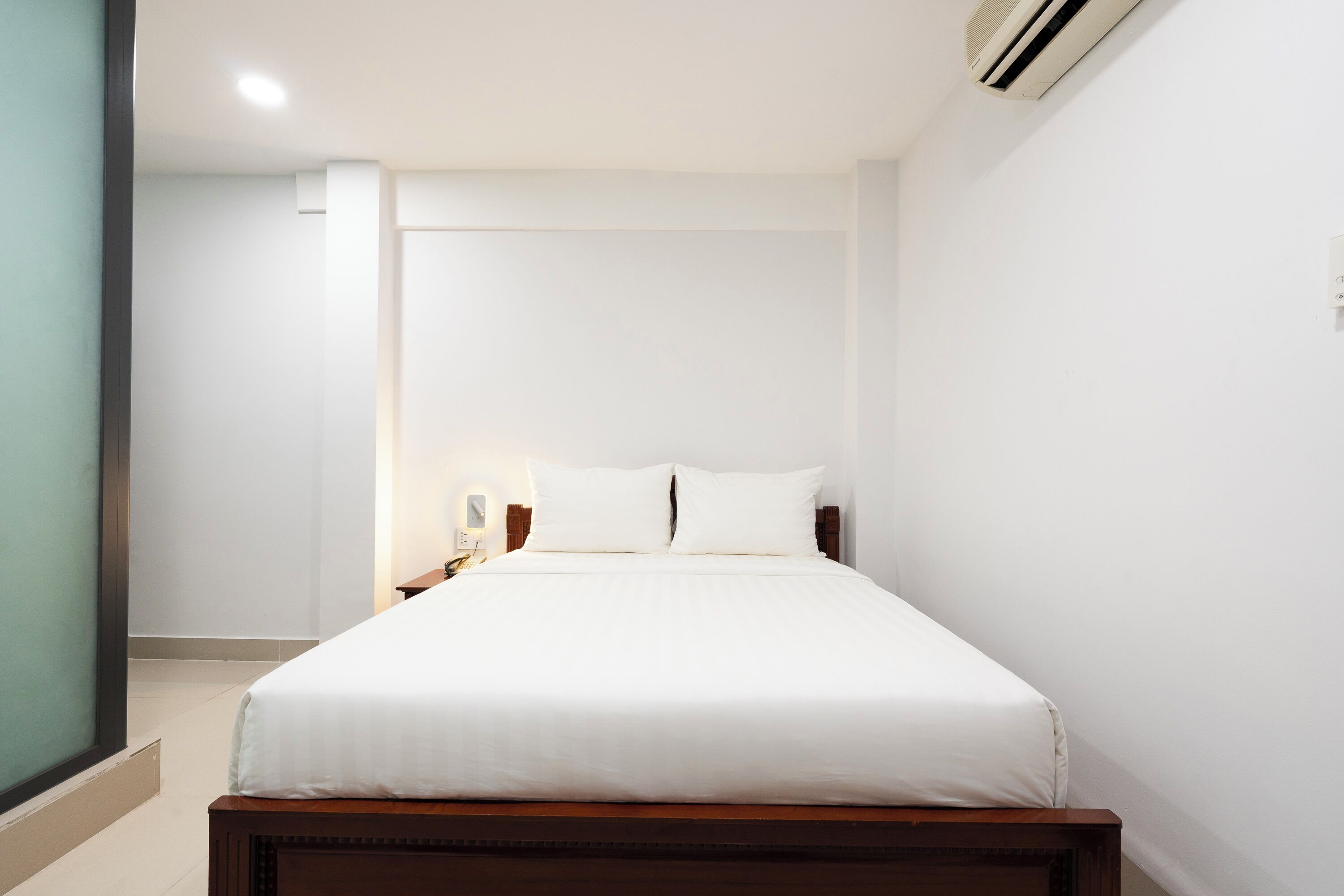 A neatly made bed in a hotel room with a wooden headboard, a nightstand with a lamp, and a wall-mounted air conditioning unit.