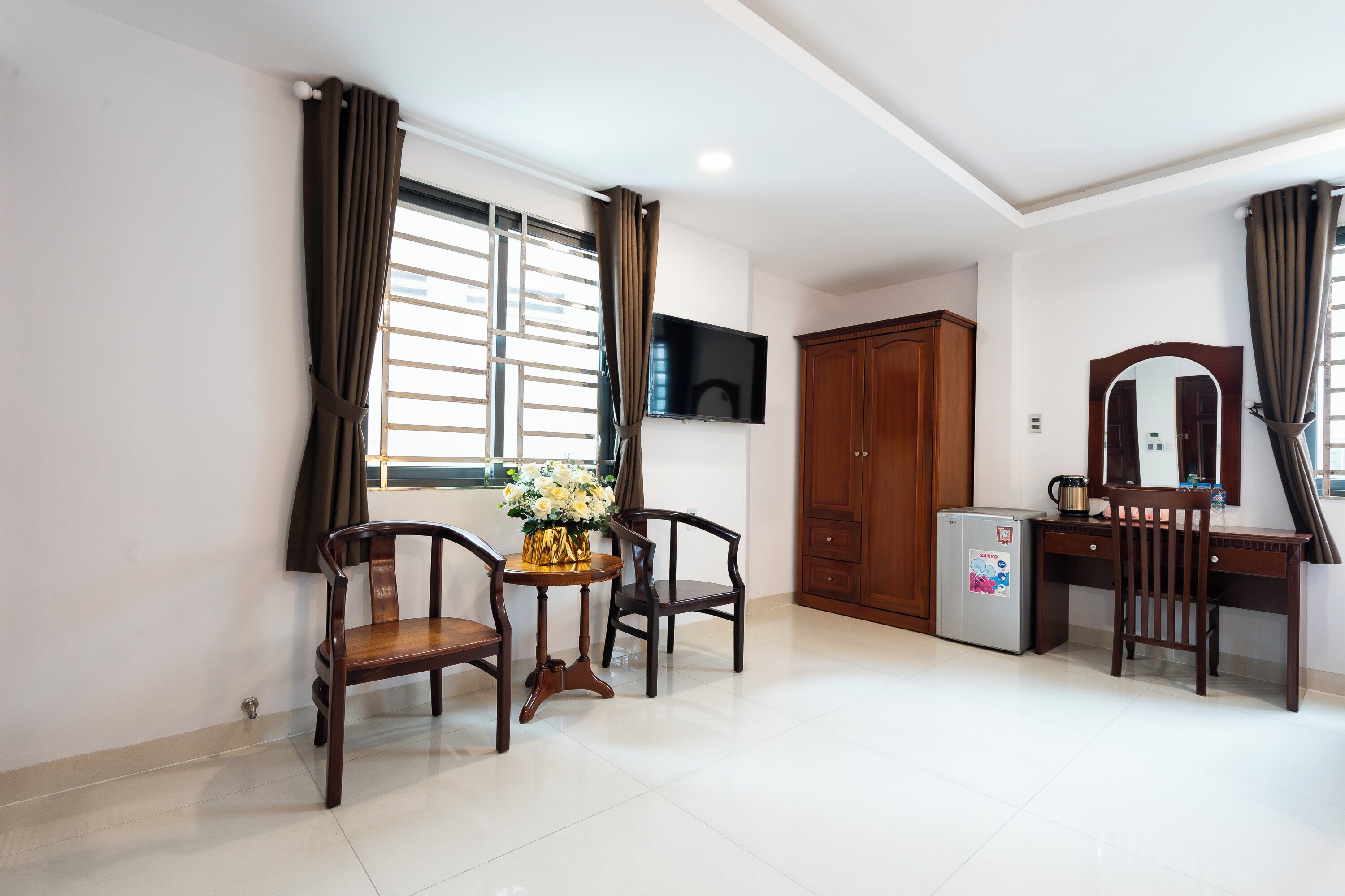 A hotel room with a wooden desk, two chairs, a small table with a vase, a refrigerator, a TV mounted on the wall, and a wardrobe.