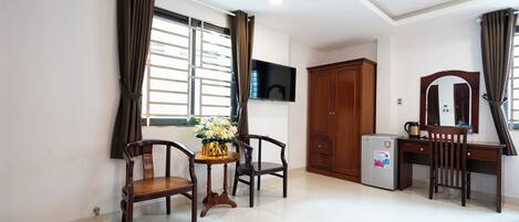 A hotel room with a wooden desk, two chairs, a small table with a vase, a refrigerator, a TV mounted on the wall, and a wardrobe.