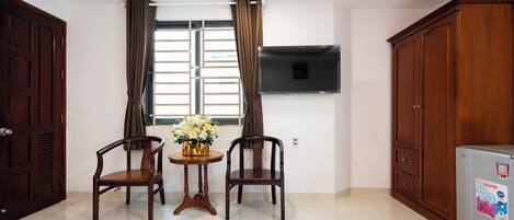 Una habitación con dos sillas de madera, una mesa pequeña con un florero, un televisor montado en la pared y un refrigerador.