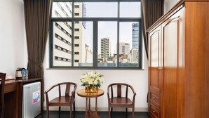 A room with a large window, two wooden chairs, a small round table with a vase of flowers, a wooden wardrobe, and a compact refrigerator. - Hoang Luxury Hotel Dist 1 Ho Chi Minh (Ho Chi Minh City)
