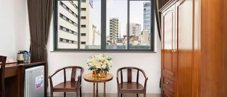 Una habitación con un ventanal grande, dos sillas de madera, una mesita redonda con un jarrón de flores, un armario de madera y un refrigerador pequeño.