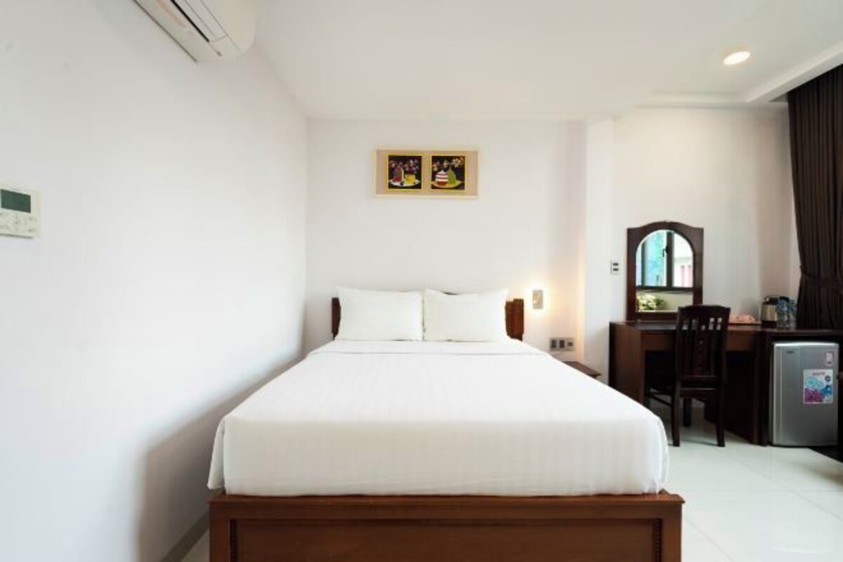 A hotel room with a single bed, a desk with a chair, a small refrigerator, and a wall-mounted mirror.