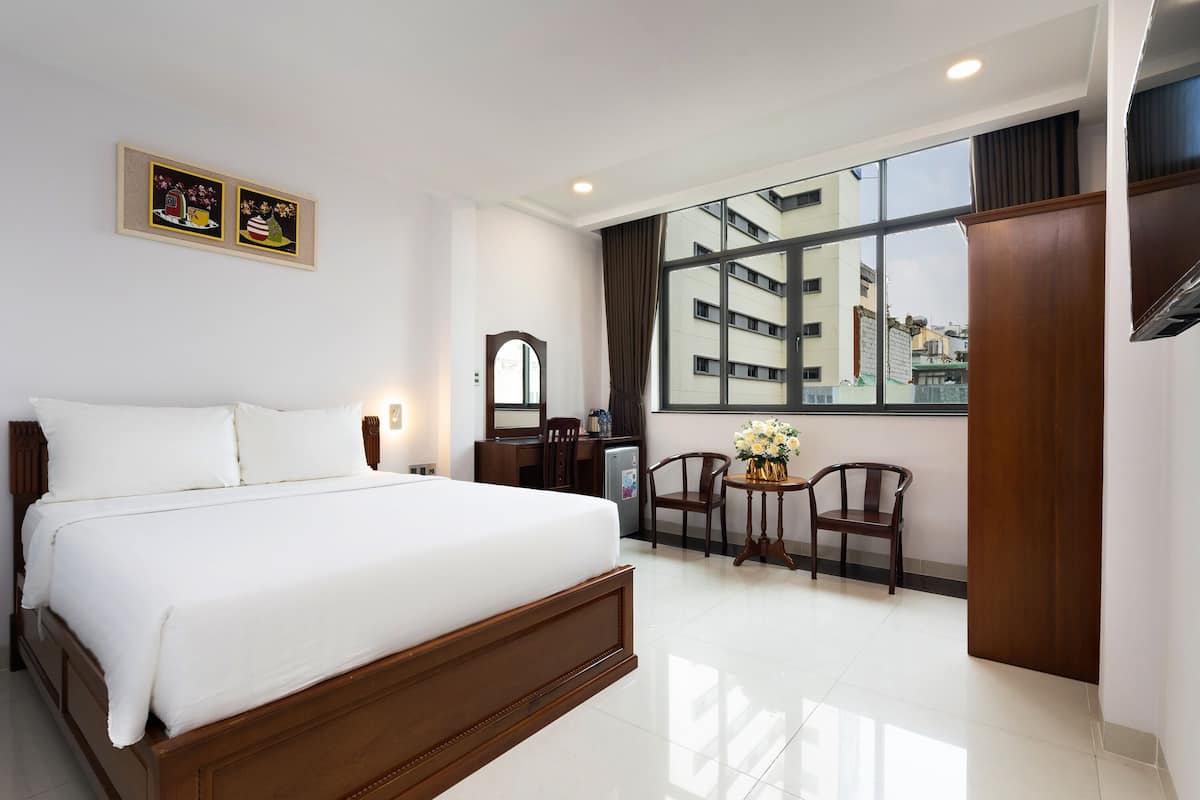 Ein Hotelzimmer mit einem großen Bett, einem Schreibtisch, einem Stuhl, einem kleinen Tisch mit Blumen und einem Fenster mit Blick auf die Stadt.