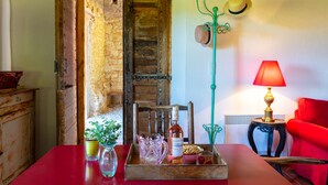 Um quarto rústico com porta de madeira, uma mesa vermelha com uma garrafa de vinho, um vaso com plantas verdes e um abajur vermelho.