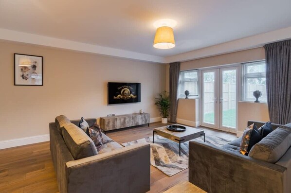 A modern living room with a sofa set, a coffee table, a TV mounted on the wall, and a large window with curtains.