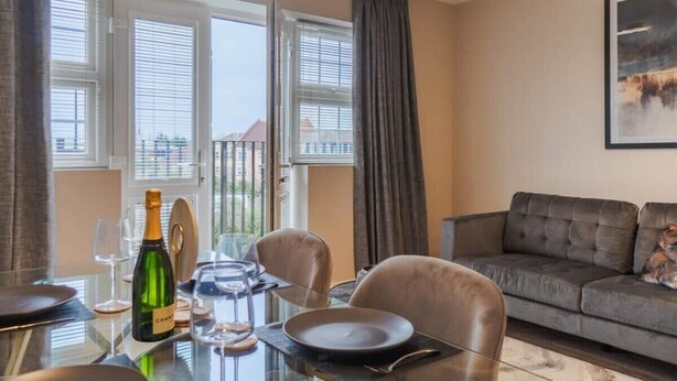 A dining area with a table set for two, a bottle of champagne, and a sofa in the background. - Castle Views| 2 Bed |Parking - VAT Q - By Tempstay (Berkshire)
