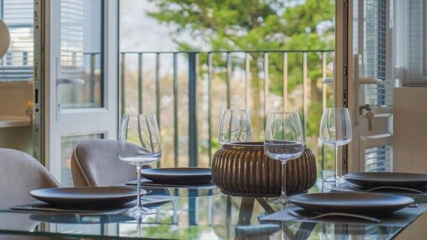 A dining table set with plates, glasses, and a decorative vase, with a view of greenery through a balcony door. - Chic 2 Bed Flat - Balcony - Parking - By Tempstay (Berkshire)
