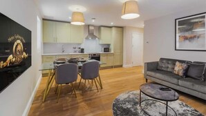A modern living room with a sofa, a dining table with chairs, a kitchen area, and pendant lighting. - 1BR Gem, Private Parking - VAT Q - By Tempstay (Berkshire)