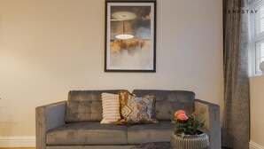 A grey sofa with patterned cushions, a framed abstract painting, a vase with flowers, and a window with curtains. - Charming 2 BDR in Windsor - VAT Q - By Tempstay (Berkshire)