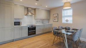 A modern kitchen with a dining area, featuring light-colored cabinets, a stainless steel range hood, a microwave, and a dining table with chairs. - Charming 2 BDR in Windsor - VAT Q - By Tempstay (Berkshire)