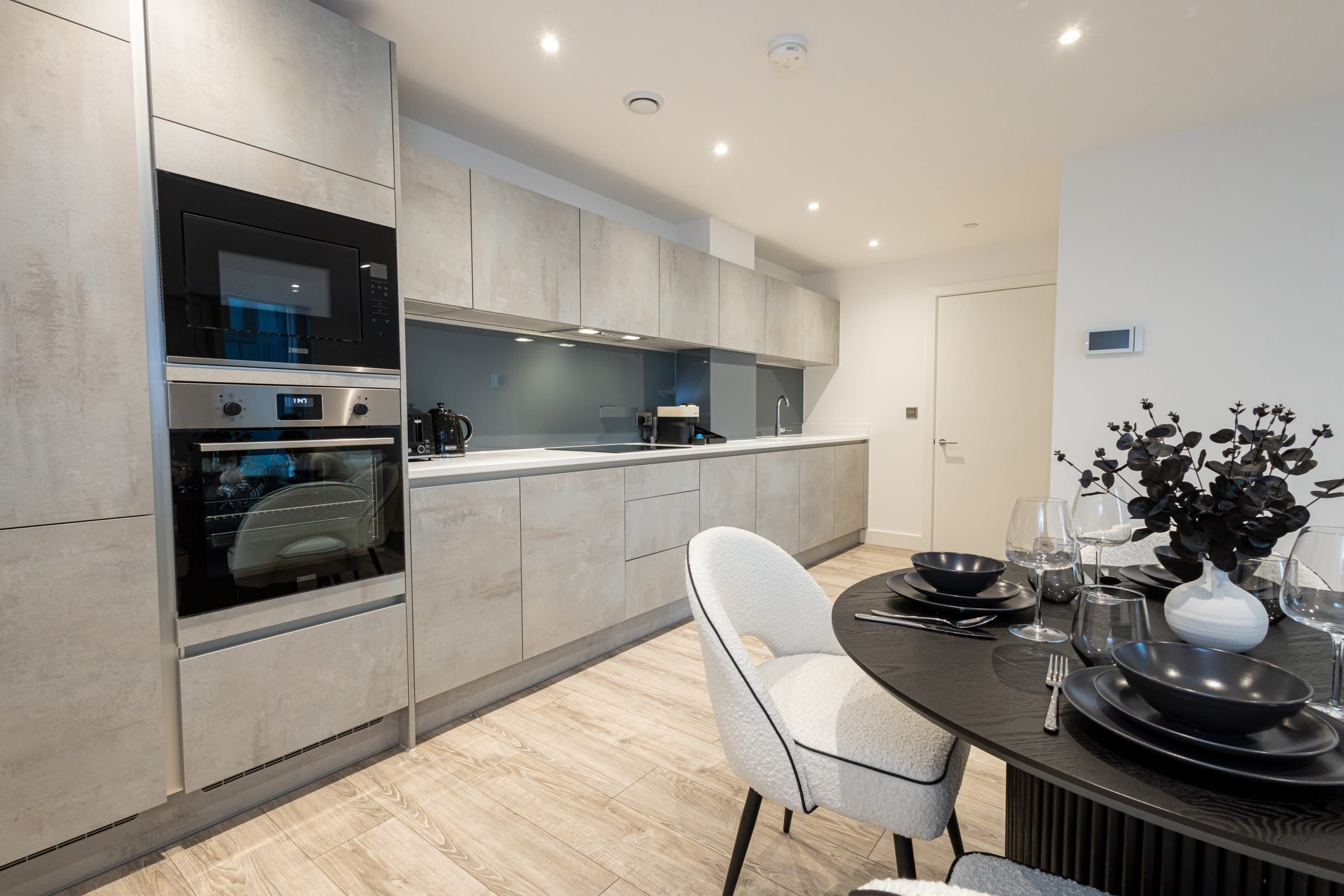 Une cuisine moderne dotée d’appareils électroménagers intégrés, un coin repas avec une table dressée pour deux personnes et une chaise blanche.