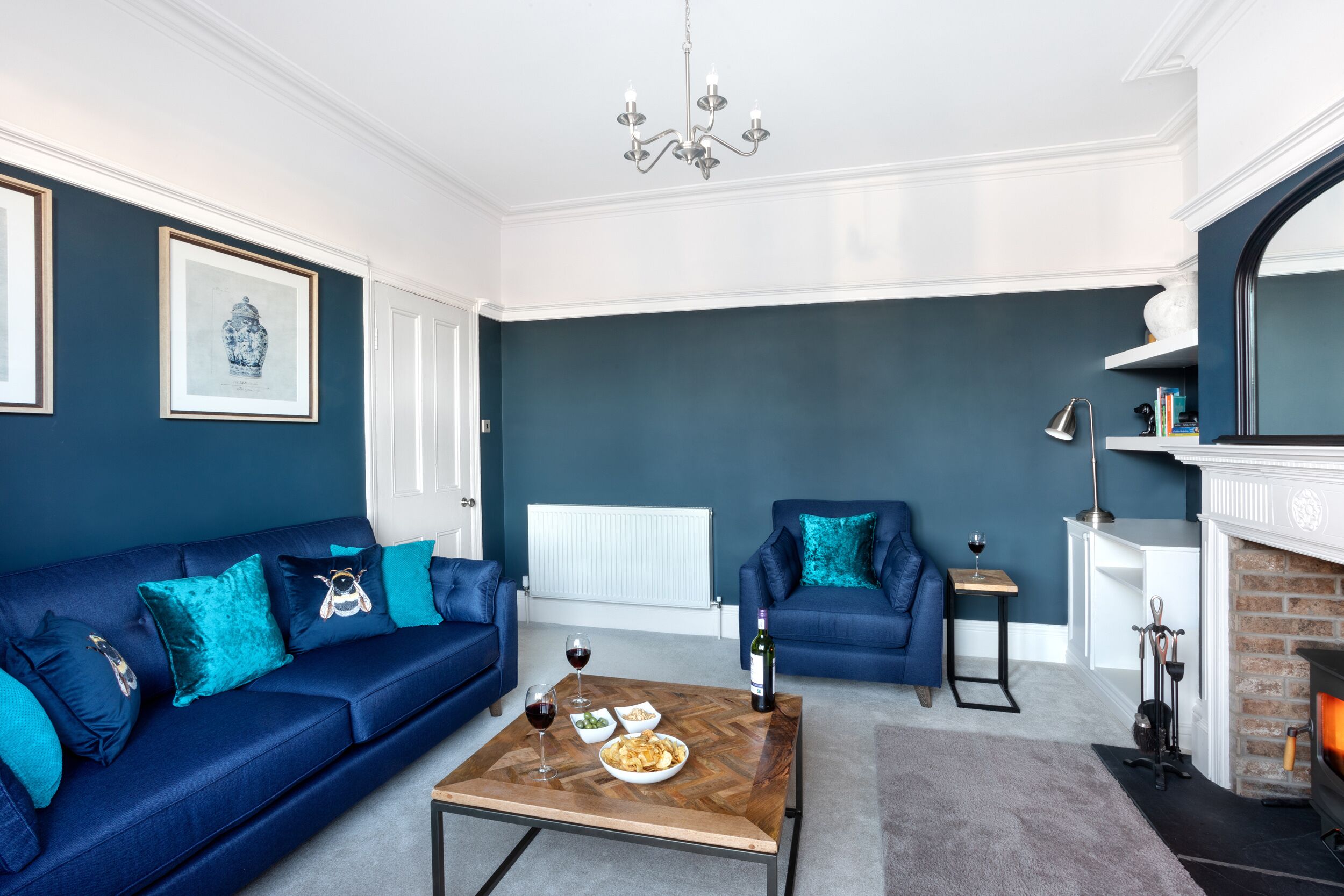 A modern living room with a blue sofa, a coffee table with food and drinks, a fireplace, and a chandelier.