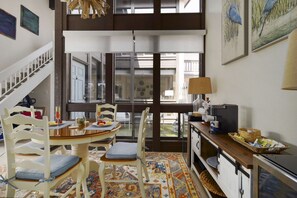 Un coin repas avec une table ronde, des chaises et un tapis à motifs. Un escalier se trouve à gauche, et à droite, un espace cuisine équipé d’appareils électroménagers et d’une machine à café.