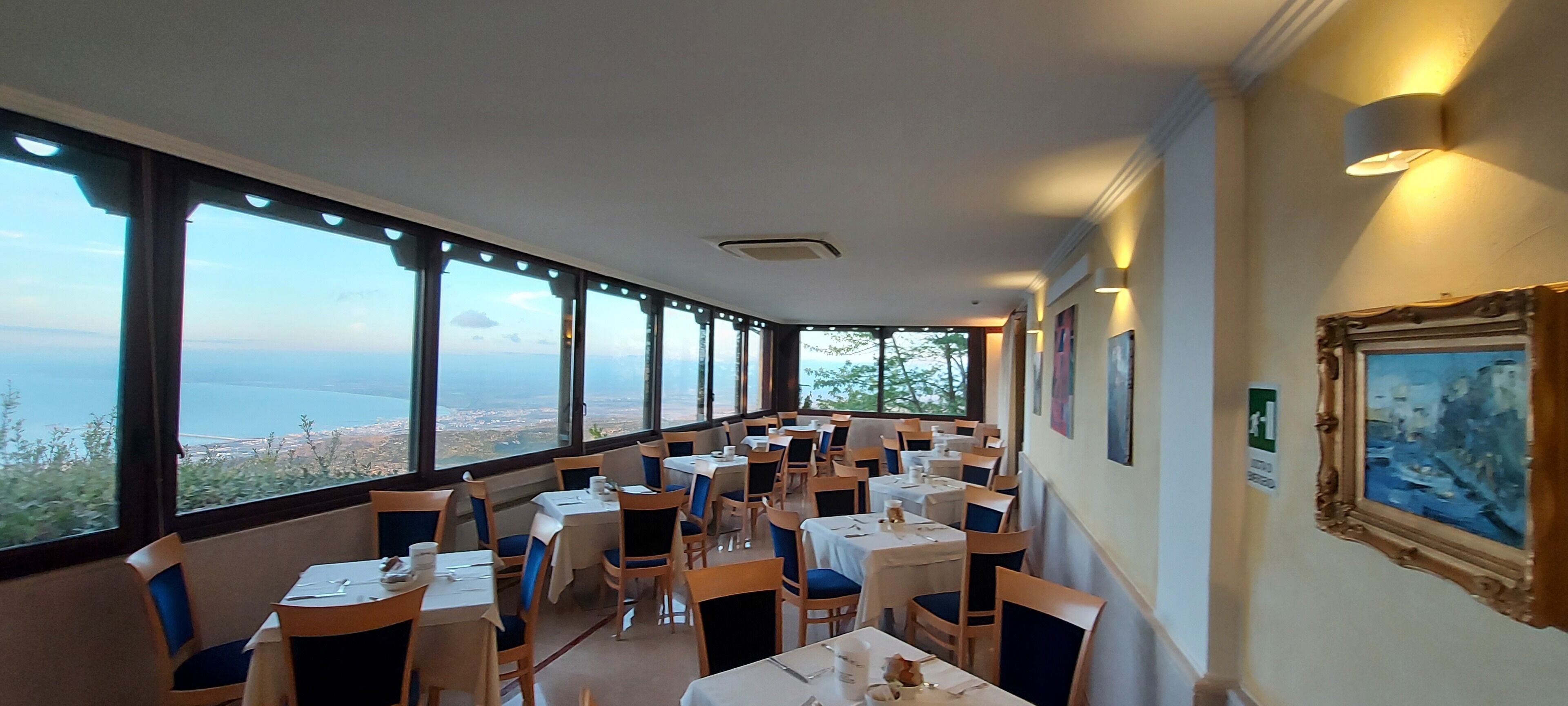A restaurant with tables and chairs arranged for dining, large windows offering a view of the sea, and framed artwork on the walls.