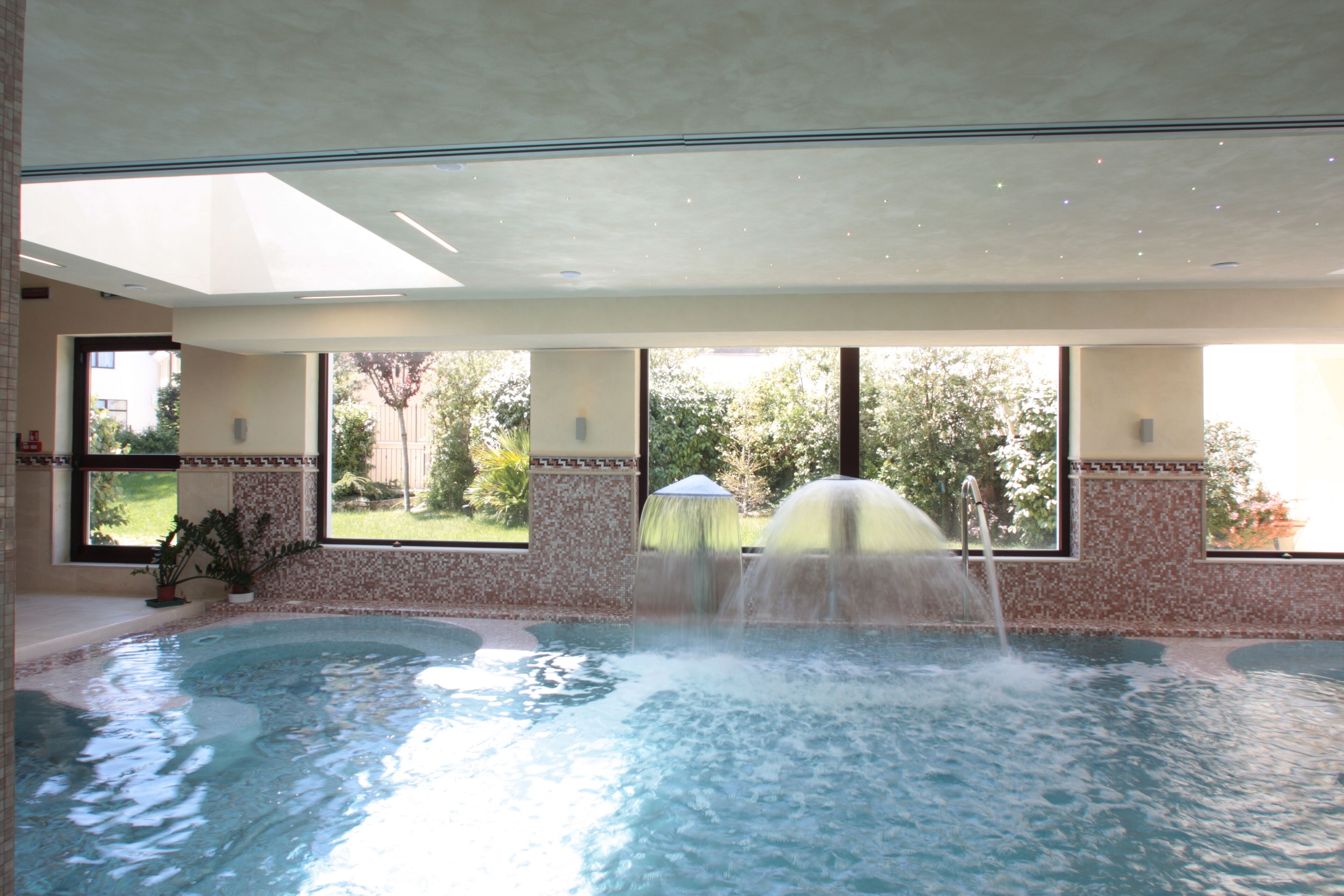 An indoor swimming pool with a water fountain, large windows, and a view of greenery outside.