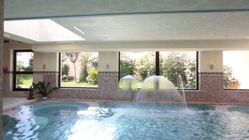 An indoor swimming pool with a water fountain, large windows, and a view of greenery outside.