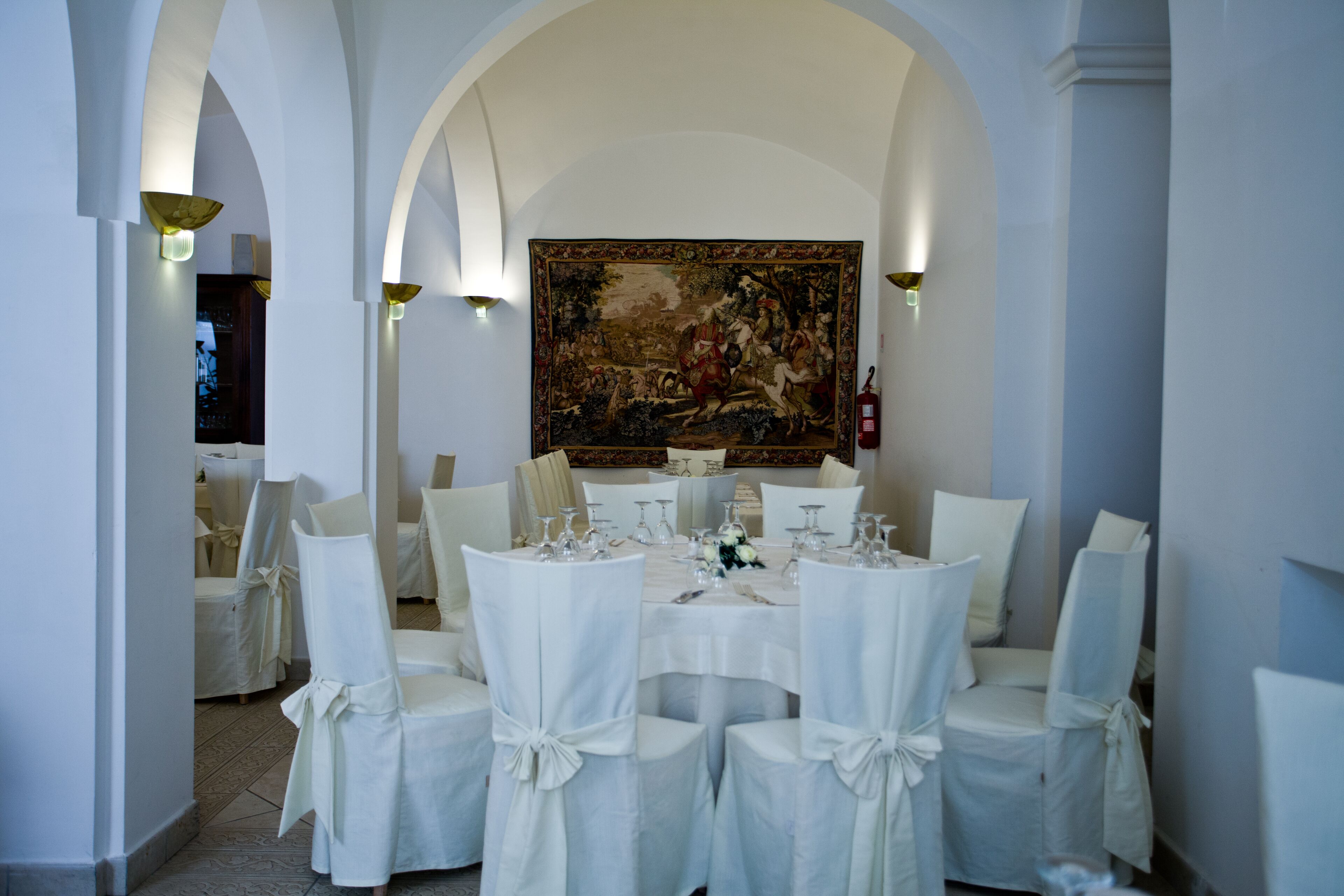 A formal dining room with white chairs and tables, a large classical painting on the wall, and a chandelier.