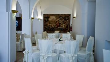 A formal dining room with white chairs and tables, a large classical painting on the wall, and a chandelier.