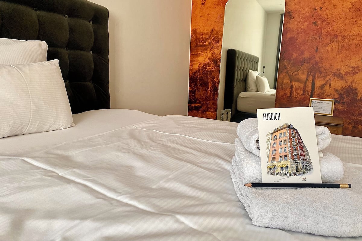 A neatly made bed with white linens, a black headboard, and a folded towel with a 'FÜRDI' card and a pencil on it.