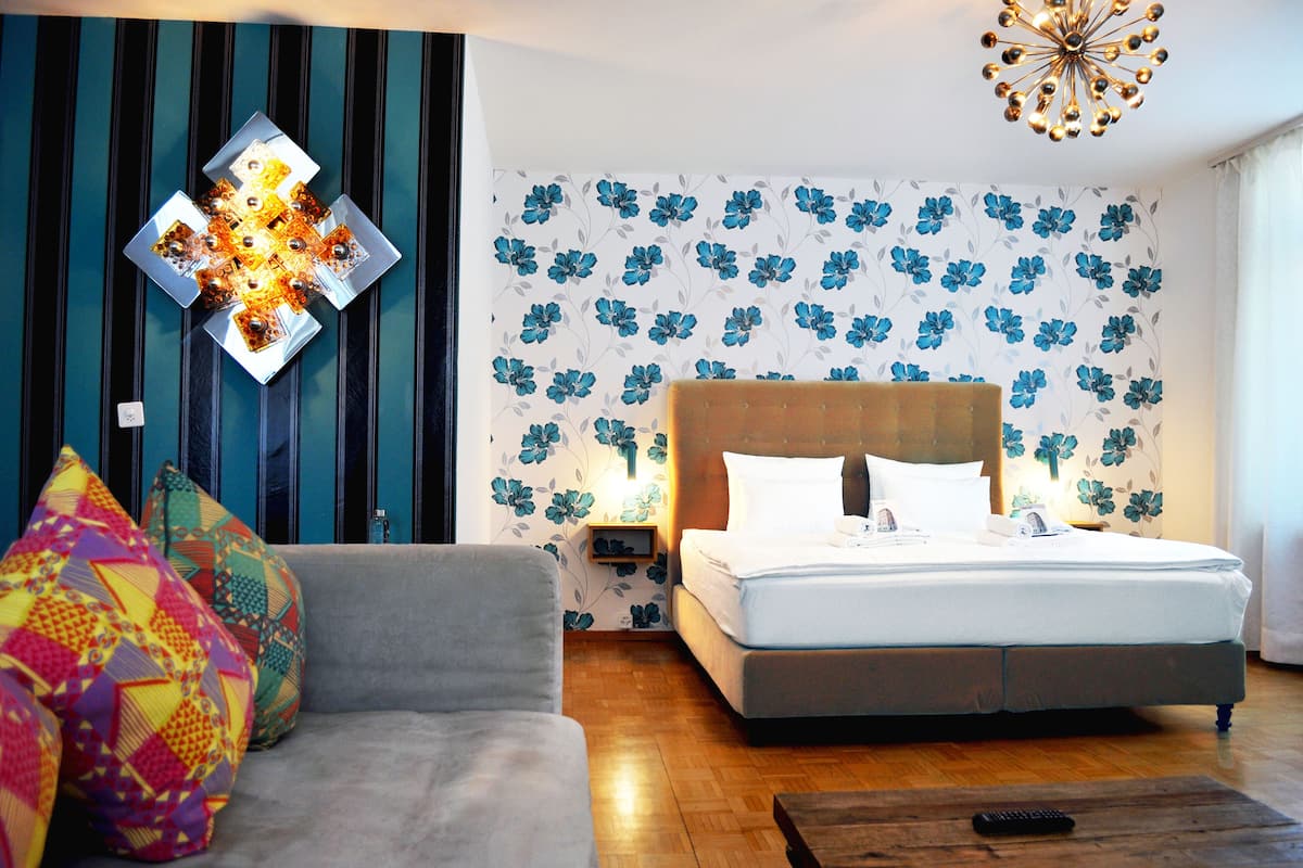 A bedroom with a bed, a sofa, colorful pillows, a wall with a floral pattern, and a unique light fixture.