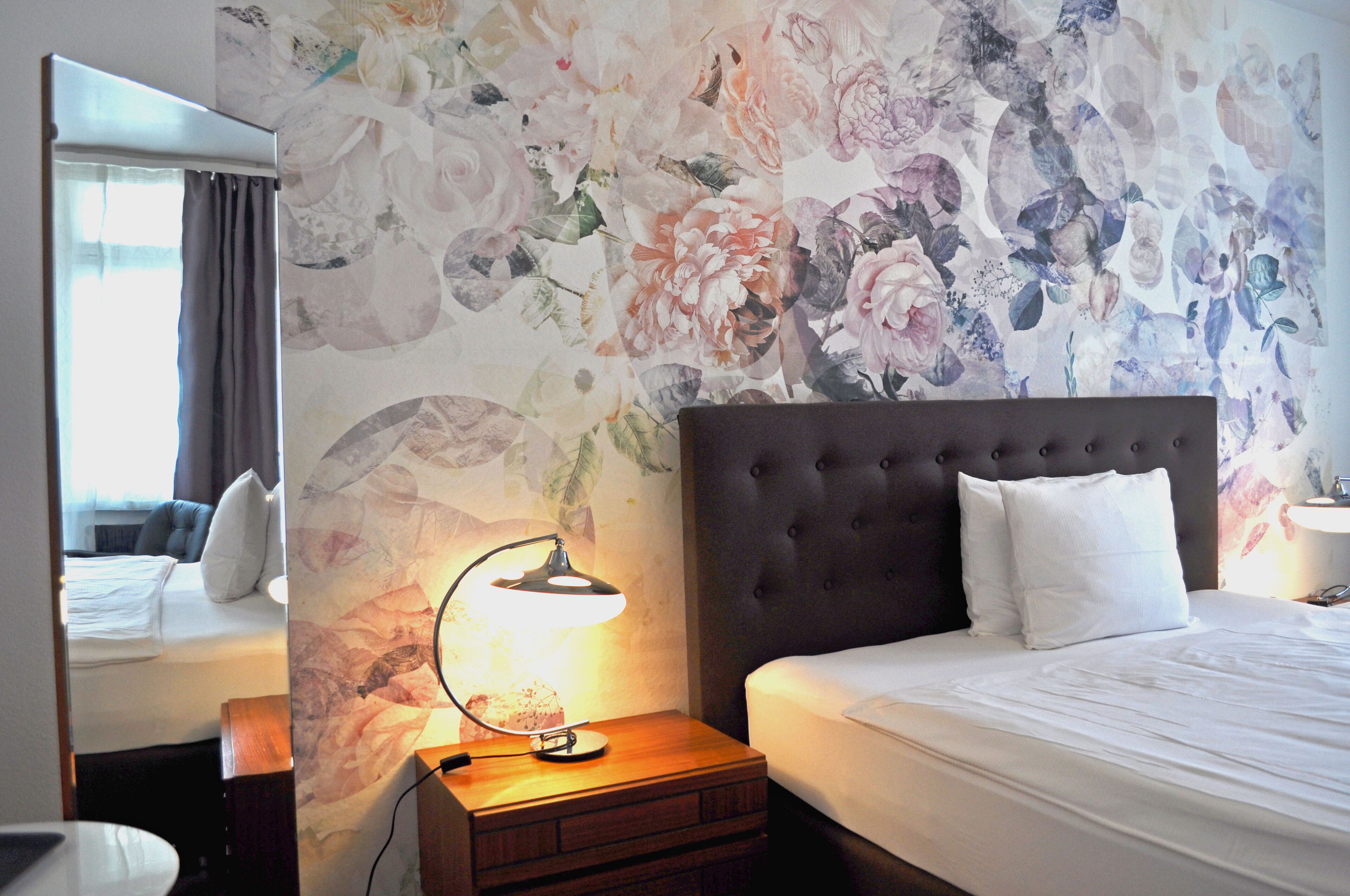 A hotel room with a large bed, a nightstand with a lamp, a mirror, and a floral wallpaper.