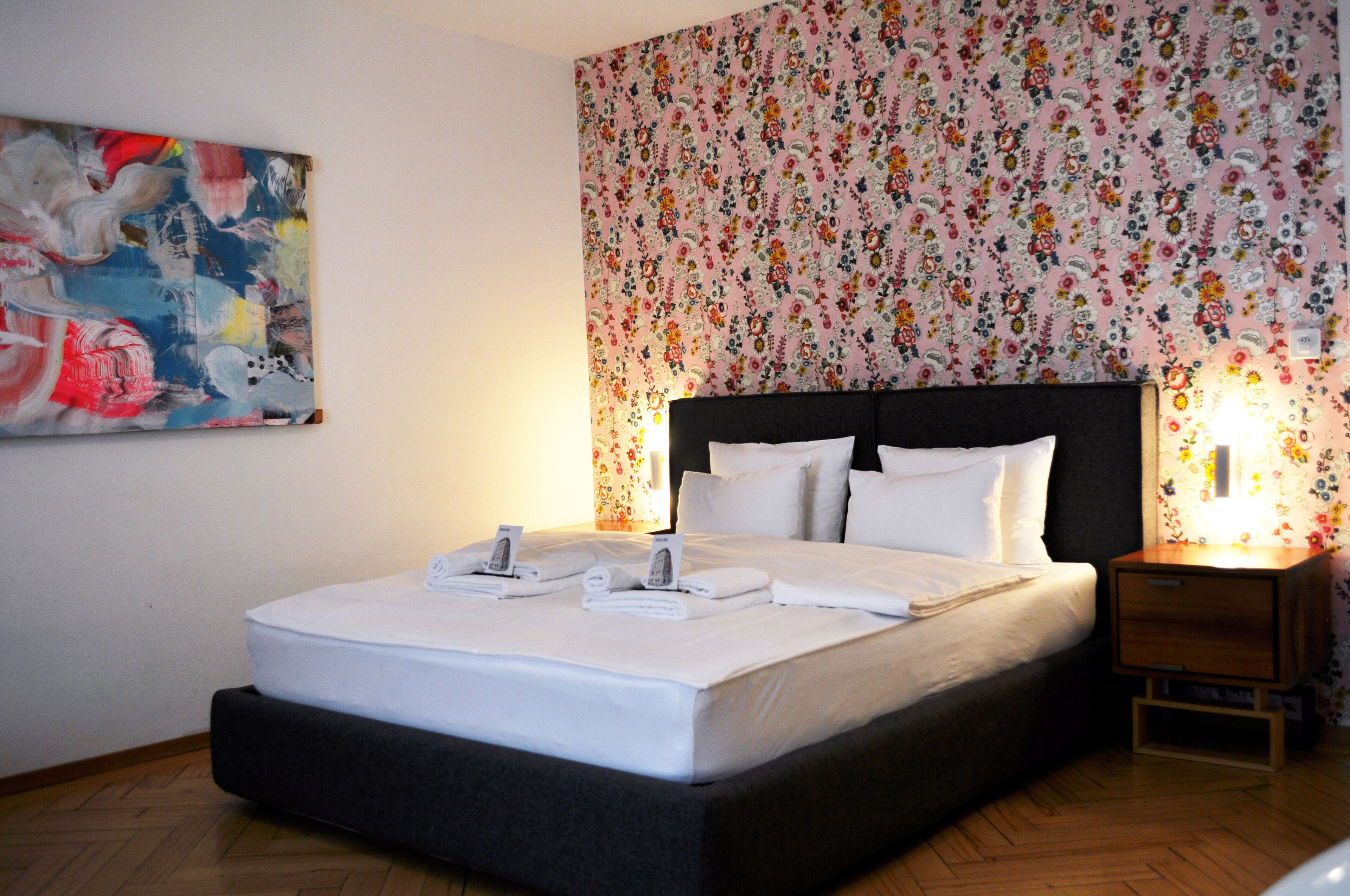 Una cama bien hecha con sábanas y toallas blancas, un papel tapiz floral y un cuadro abstracto moderno en la pared.