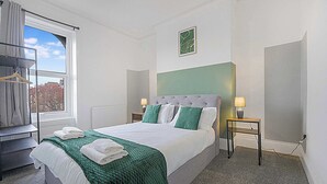Hotellihuone, jossa on sänky, ikkuna, patteri, yöpöytä lampun kanssa ja naulakko.
