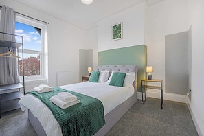 Ein Hotelzimmer mit Bett, Fenster, Heizkörper, Nachttisch mit Lampe und Kleiderhaken.