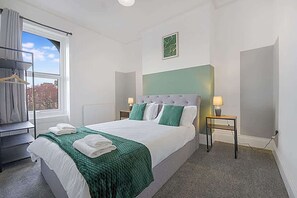Habitación de hotel con cama, ventana con vistas, mesita de noche con lámpara y perchero.
